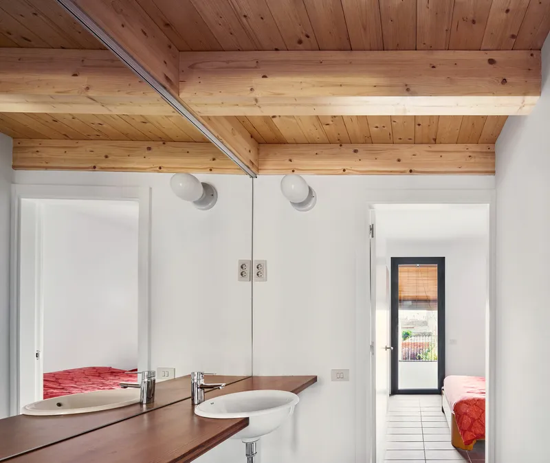 Bathroom with timber vanity and exposed ceiling joists looking through doorways toward bedrooms with red bedding