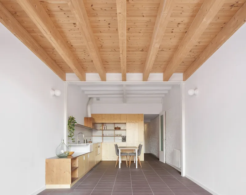 Open-plan kitchen and dining space with exposed timber beams, plywood cabinetry, and terracotta floor tiles