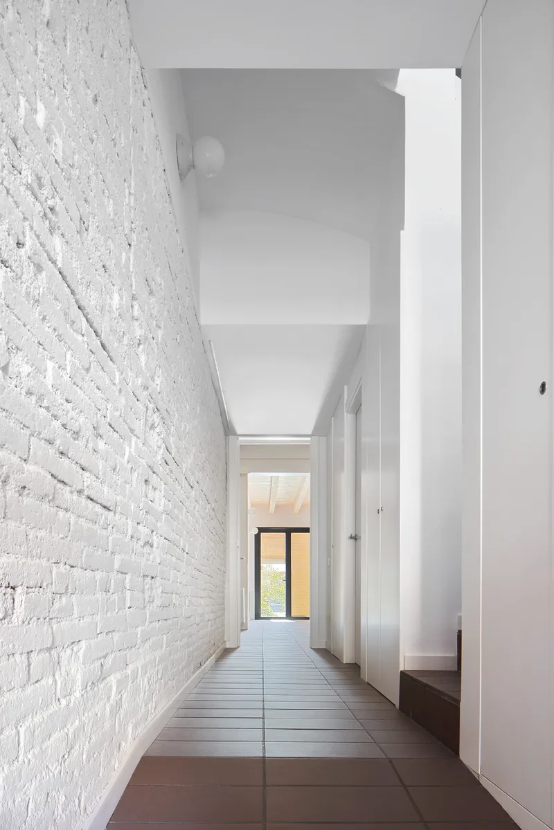 Narrow corridor with painted white brick wall and terracotta tile floor leading to a distant window