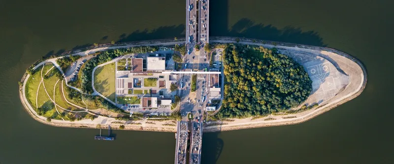 Top-down view of the oval island showing low-rise volumes surrounded by planted areas and bridge connections
