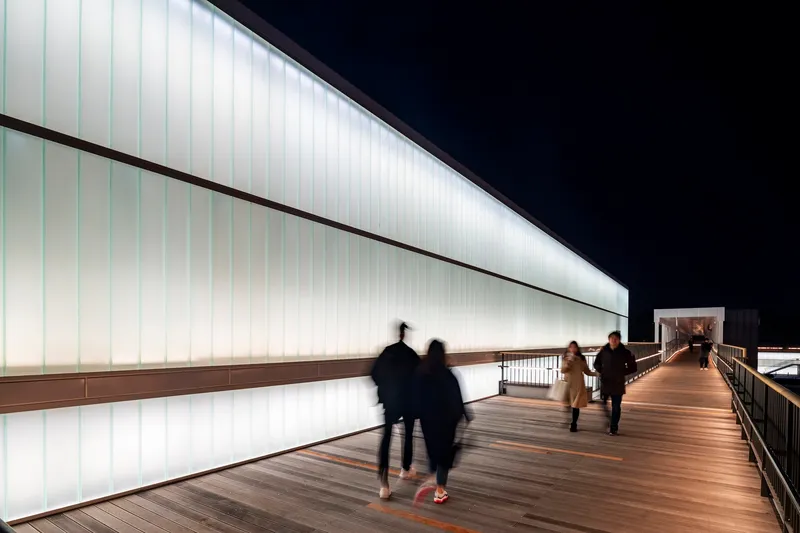 Elevated timber walkway along a backlit translucent panel wall with blurred figures at night