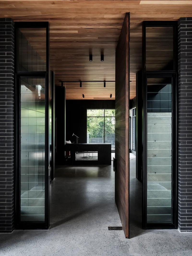 Entry hallway with timber-clad ceiling and pivot door flanked by glass panels with dark brick walls