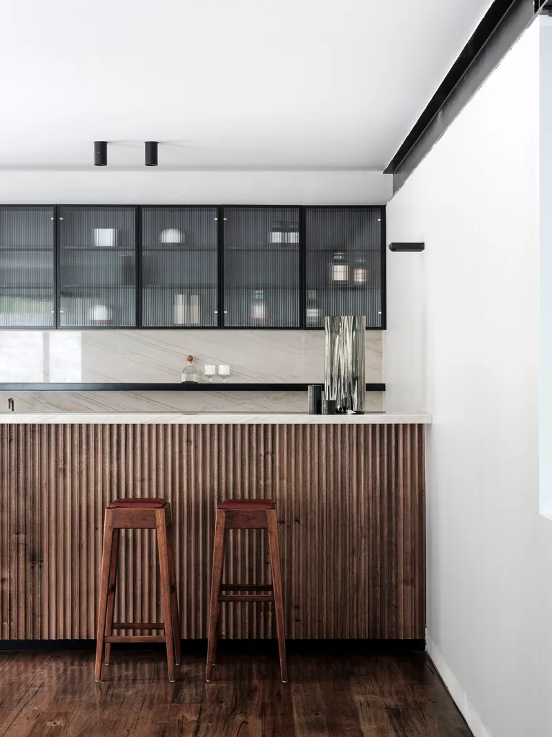 Kitchen island with vertical timber slat cladding and two wooden stools beneath reeded glass cabinets
