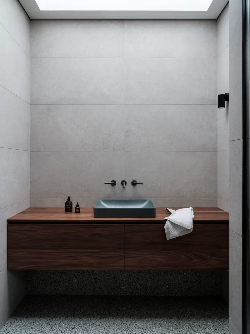 Bathroom vanity with floating walnut cabinetry and charcoal basin below large format concrete tile walls