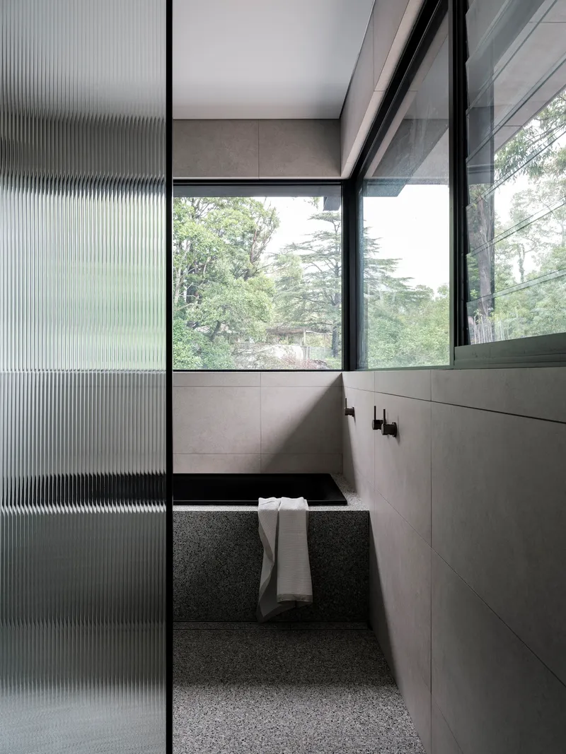 Sunken bathtub with terrazzo surround framed by fluted glass screen and windows overlooking garden