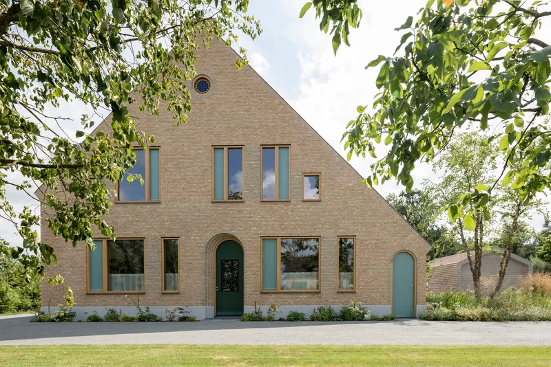 Gabled brick facade with varied window openings and arched door framed by young trees in spring
