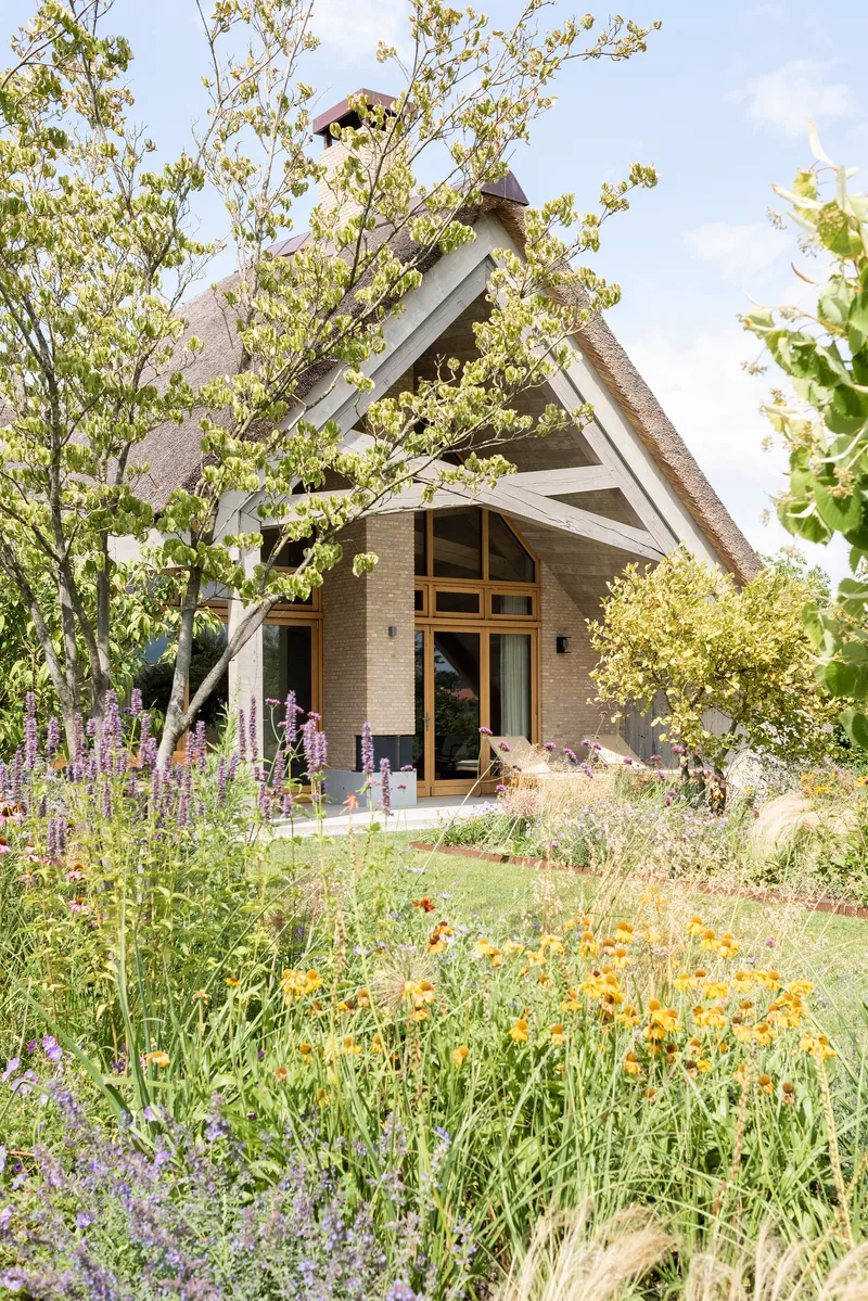 Entrance facade with thatched gable roof surrounded by flowering perennials and young trees