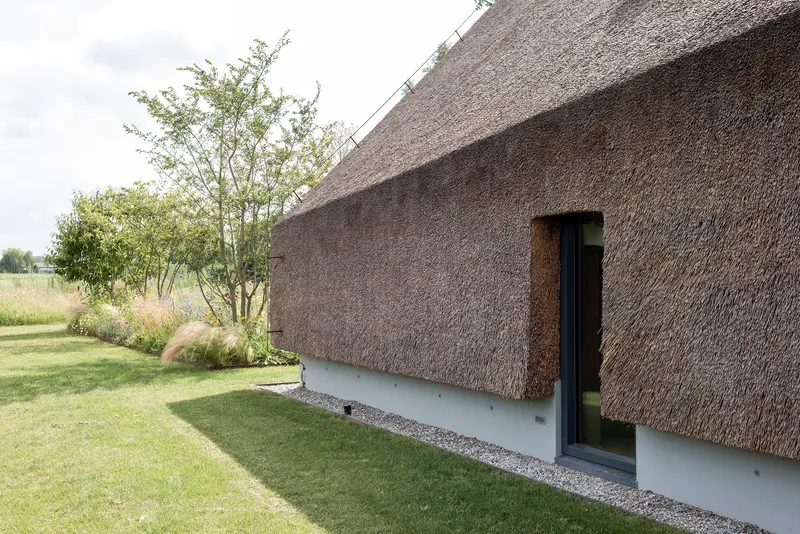 Thatched roof extending down to a white plinth with a narrow glazed door and gravel path