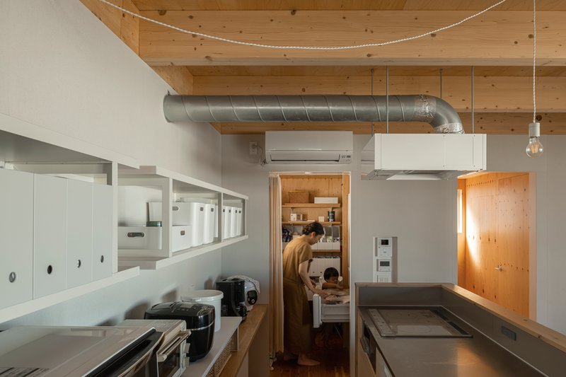 Kitchen workspace with overhead exposed duct, white storage boxes, and figures visible through pantry doorway