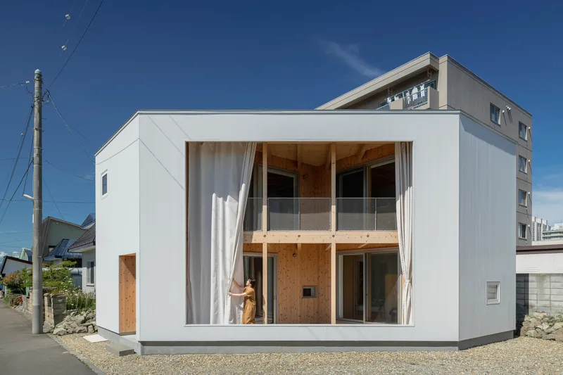 White box facade with floor-to-ceiling glazing and sheer curtains pulled back by a figure inside