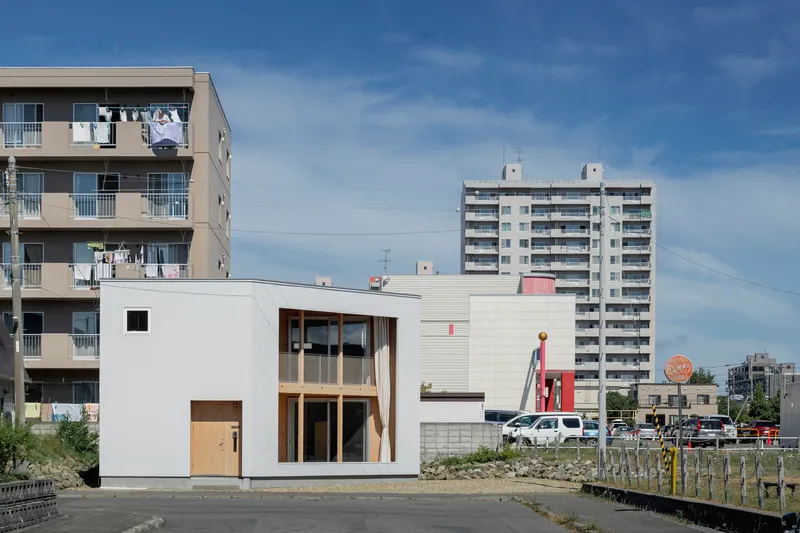 Street view of the white metal-clad volumes against mid-rise apartment buildings under blue sky