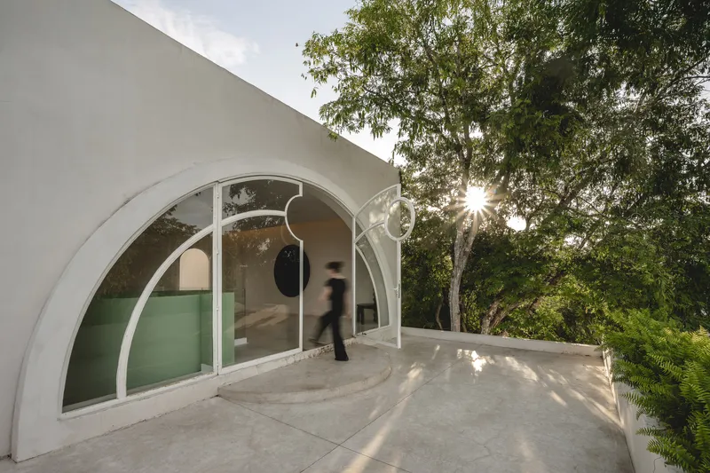 Curved glass facade with white frame opening onto stone terrace as figure passes through at sunset