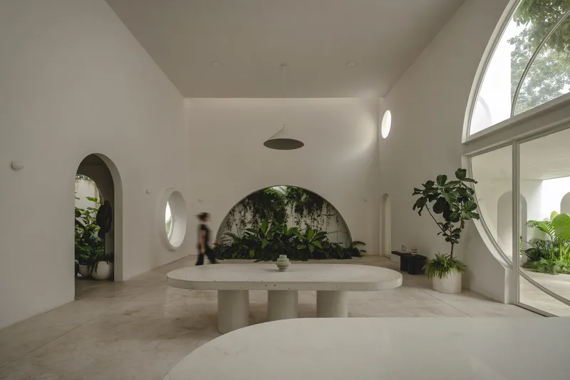 Interior room with white vaulted ceiling, circular table, arched openings, and planted alcove with blurred figure passing through