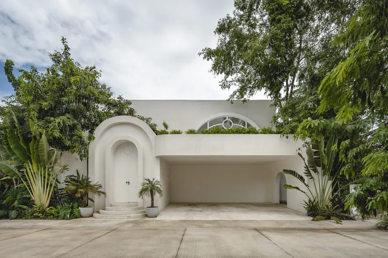 White facade with arched entrance, recessed garage, and circular window framed by banana plants and tropical trees