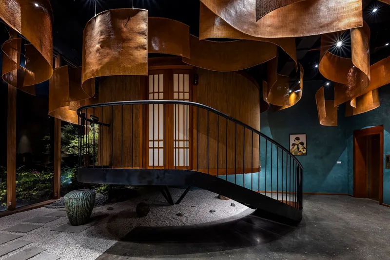 Curved staircase with black steel railings rising from a raked gravel garden bed beneath woven copper ceiling panels