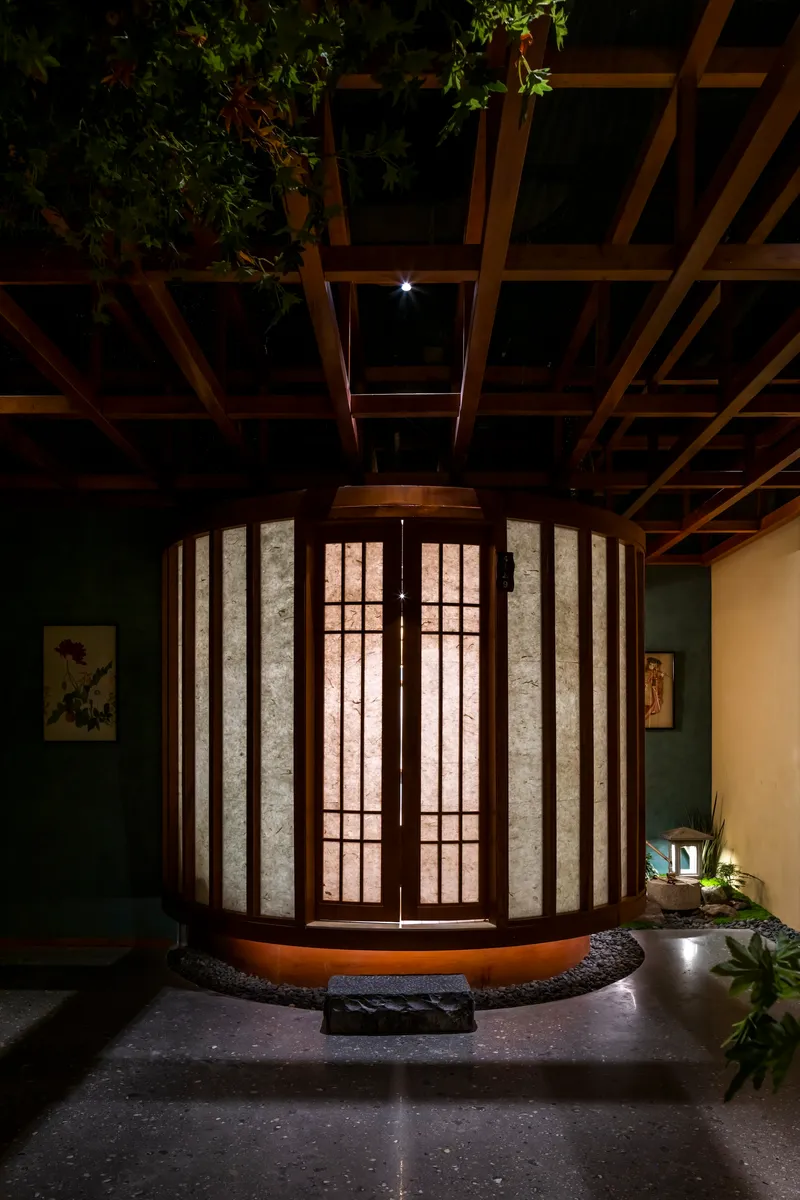 Cylindrical entry pavilion with timber frame and translucent paper panels beneath an exposed wood beam ceiling at night