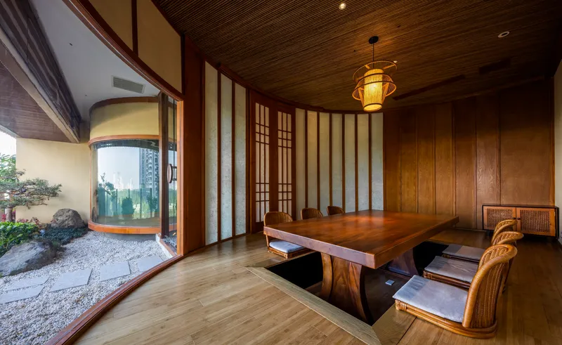 Timber dining table beside curved glass walls overlooking a small rock garden with raked gravel