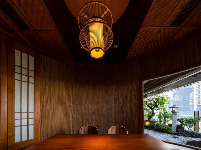 Private dining room with textured woven wall panels, bamboo lantern and opening to planted courtyard beyond