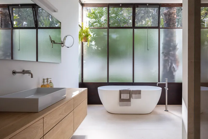 Bathroom with freestanding tub beneath tall windows with translucent lower panels and planted courtyard views