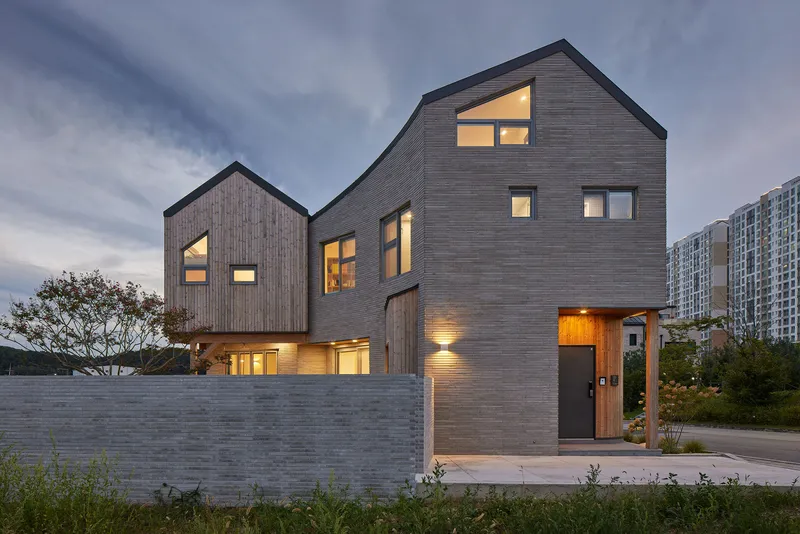 Street facade of stacked gabled volumes with dark timber cladding and illuminated windows at dusk