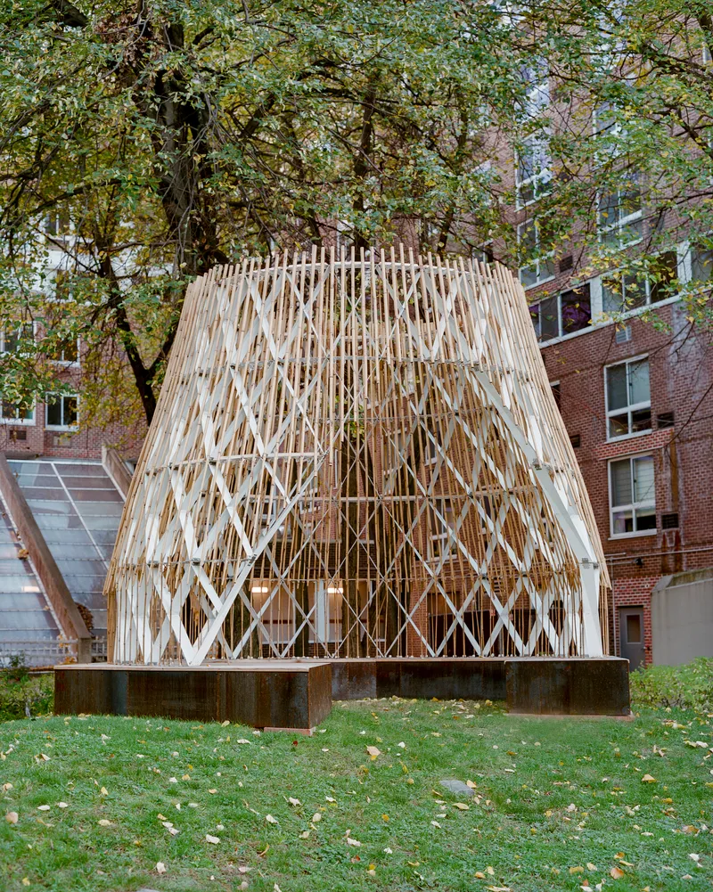Timber lattice pavilion with diamond-pattern screening on a concrete plinth in a courtyard beneath autumn trees