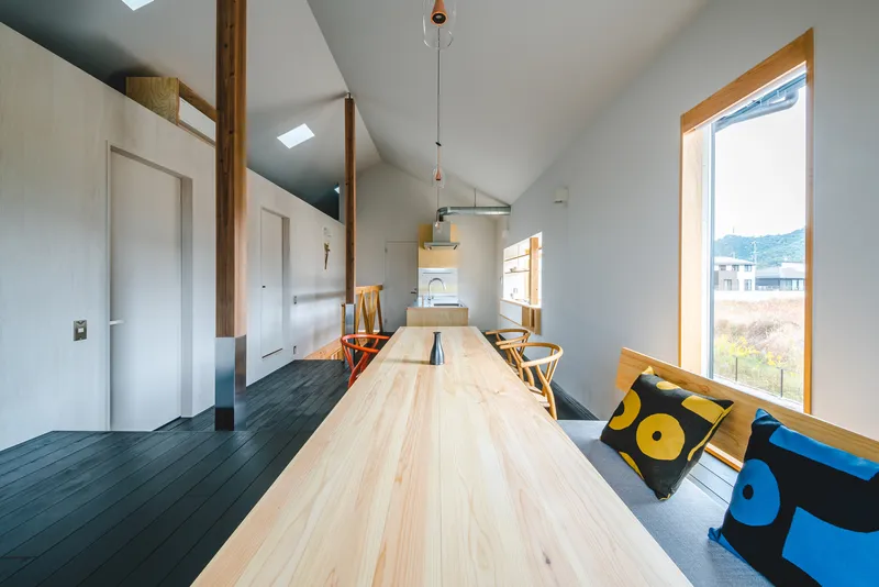 Open-plan living space with pale timber dining table extending toward the kitchen beneath a vaulted ceiling