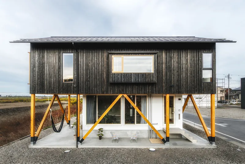 Elevated corrugated metal volume above an open ground floor with yellow diagonal bracing under overcast skies