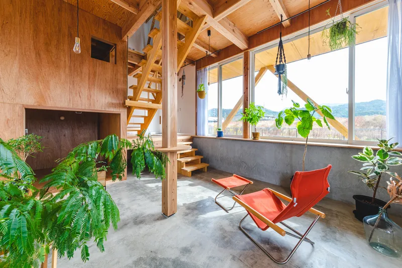 Double-height interior with exposed timber framing, concrete floor, hanging plants, and red lounge chairs