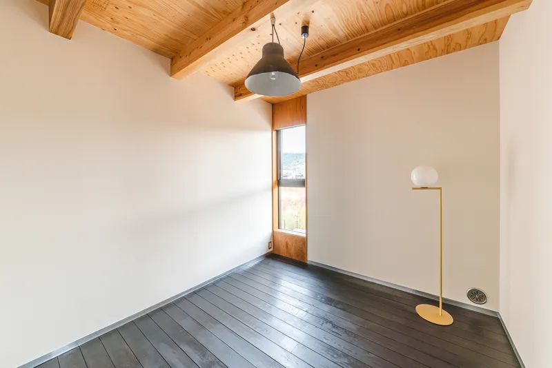 Empty room with exposed timber ceiling beams and a tall narrow window framing distant hills