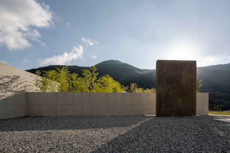 Concrete perimeter wall with freestanding textured tower on gravel surface beneath hillside in late afternoon light