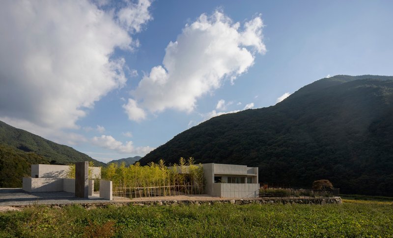 Low-rise concrete volumes with bamboo grove beside a stone retaining wall and forested hillside