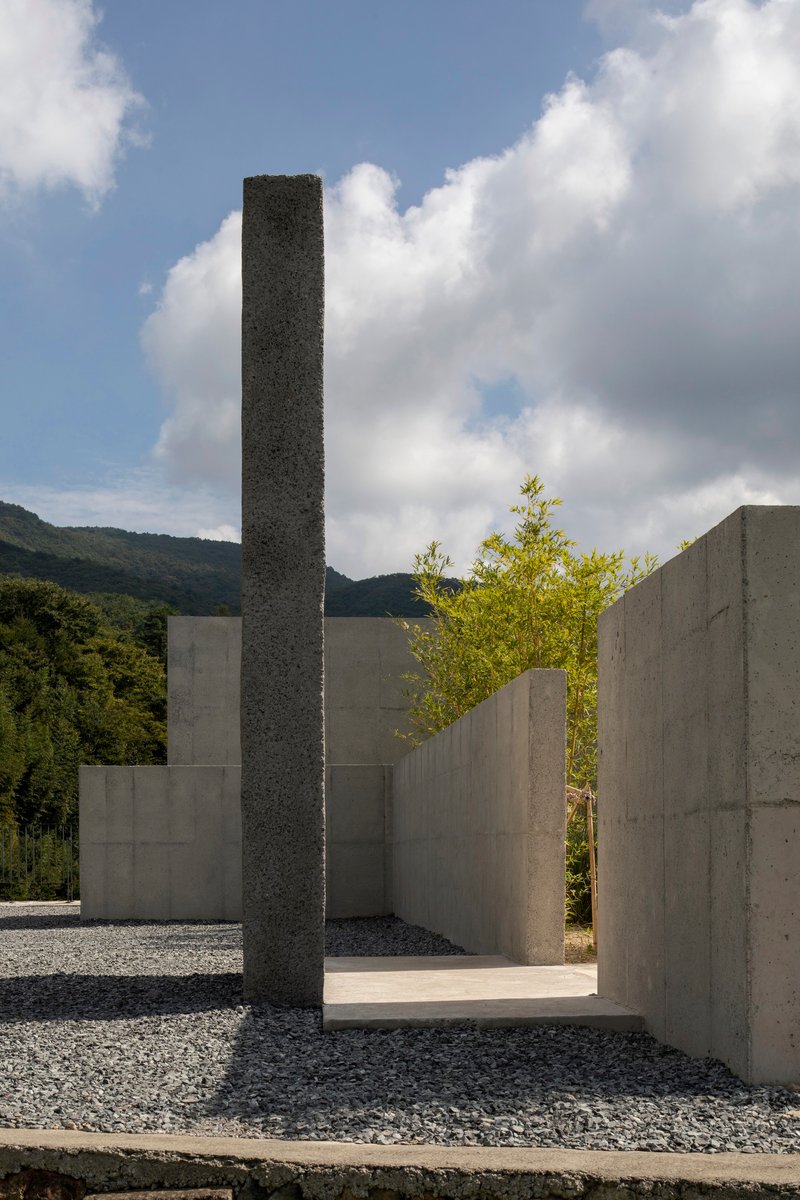 Dark aggregate pillar rising among pale concrete walls and gravel courtyard with bamboo beyond