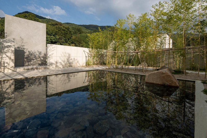 Reflecting pool mirroring white concrete walls and bamboo grove beneath clouded mountain slopes