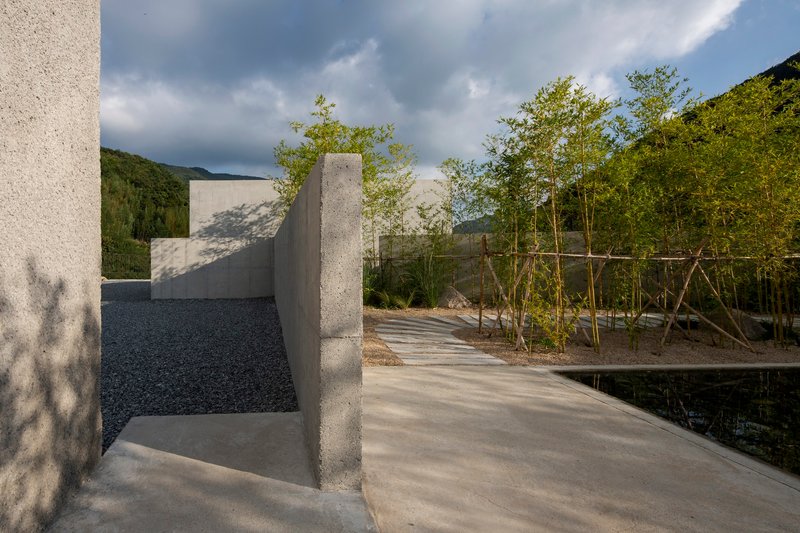 Concrete pathway curving around reflecting pool with bamboo supports and gravel beds in dappled sunlight