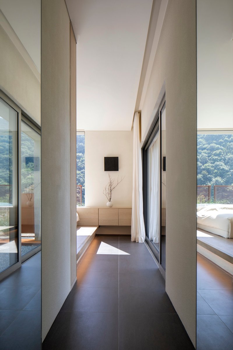 Glazed hallway passage between concrete walls with daylight entering from bedroom and terrace beyond