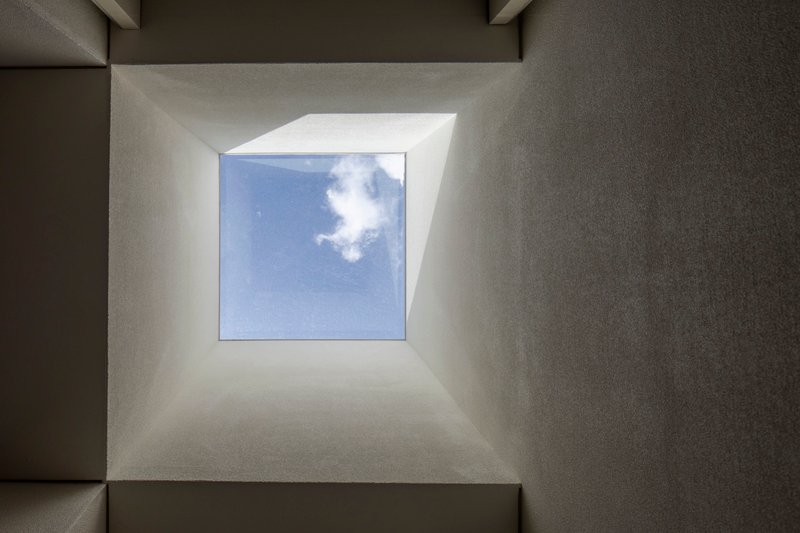 Upward view into a square skylight framed by sloped plaster walls with white clouds overhead