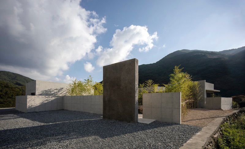 Gravel courtyard with freestanding concrete walls and tower-like element set against forested mountains