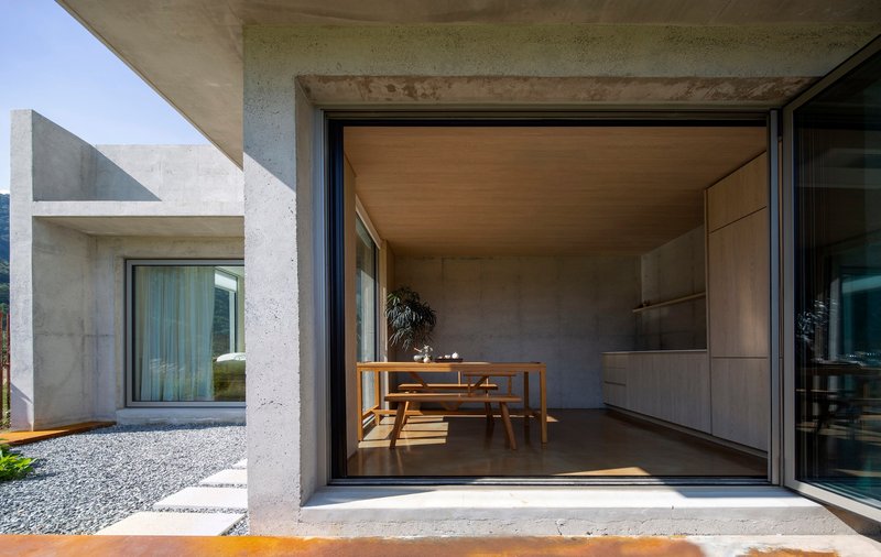 Glazed opening into interior dining area with wooden table and concrete-clad kitchen under afternoon sunlight
