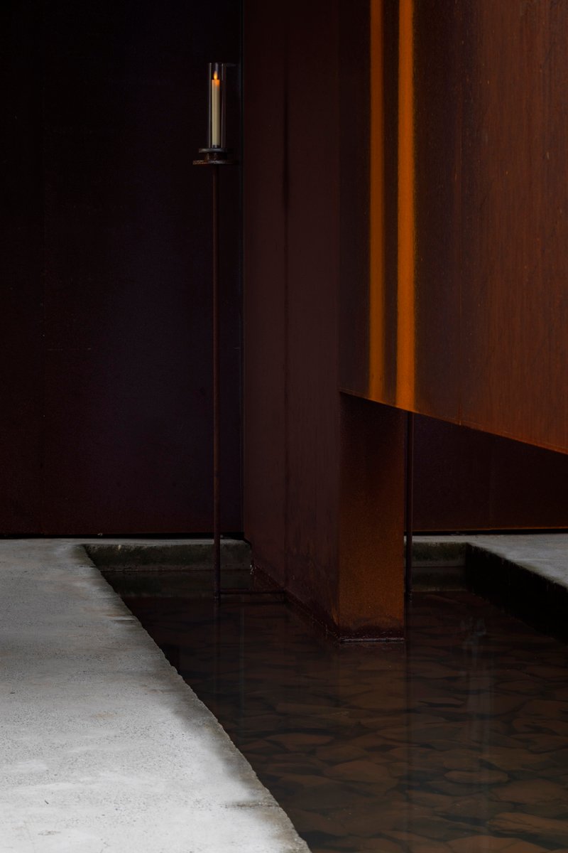 Detail of a weathered steel panel meeting a concrete floor with a tall candle in shadow