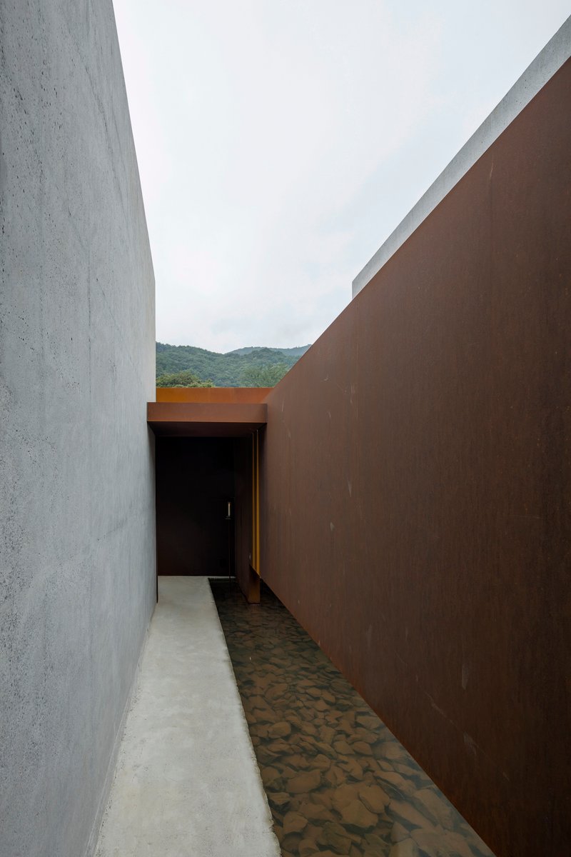 Narrow passage between concrete and weathered steel walls leading to a doorway under overcast sky