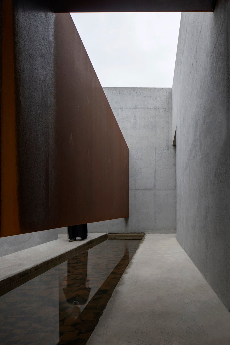 Courtyard with shallow reflecting pool between concrete walls and angled weathered steel plane overhead