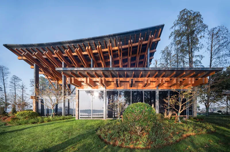 Cantilevered timber roof over stone-clad volumes with glass walls and circular planted beds at twilight