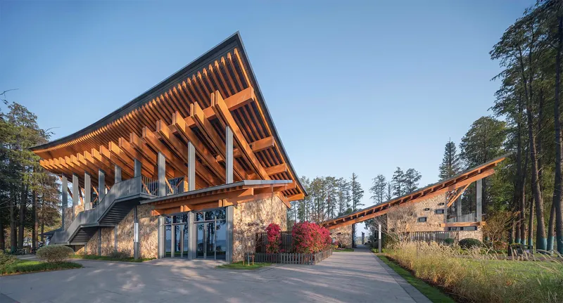 Entrance plaza with timber canopy sheltering stone-clad lower level and connecting walkway at dusk