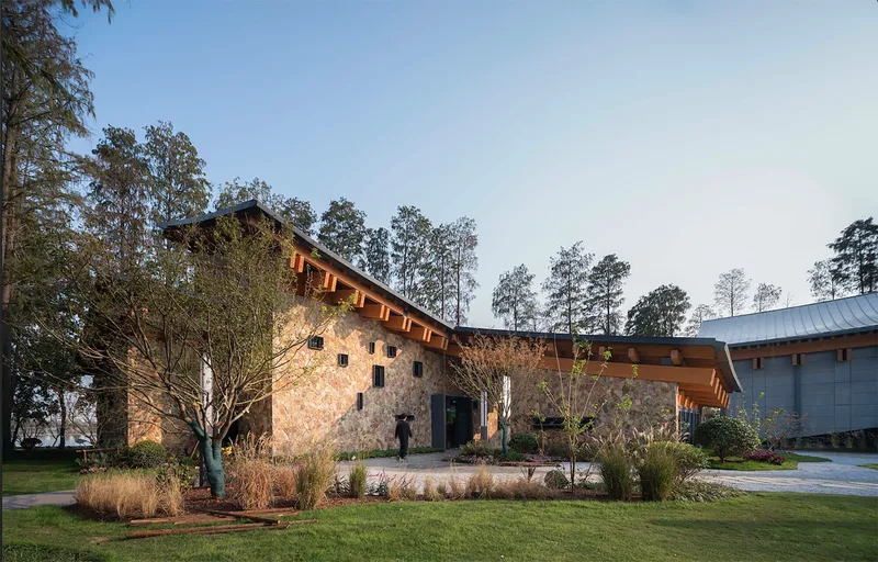 Rammed earth wall and planted beds with young trees set within mown lawn under pale sky