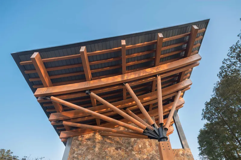 Underside view of cantilevered timber roof with radiating columns and stone base at dusk