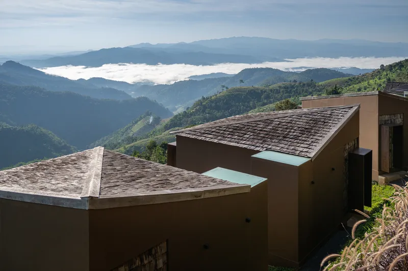 Shingled roof volumes descending a hillside with low clouds hanging over forested valleys beyond