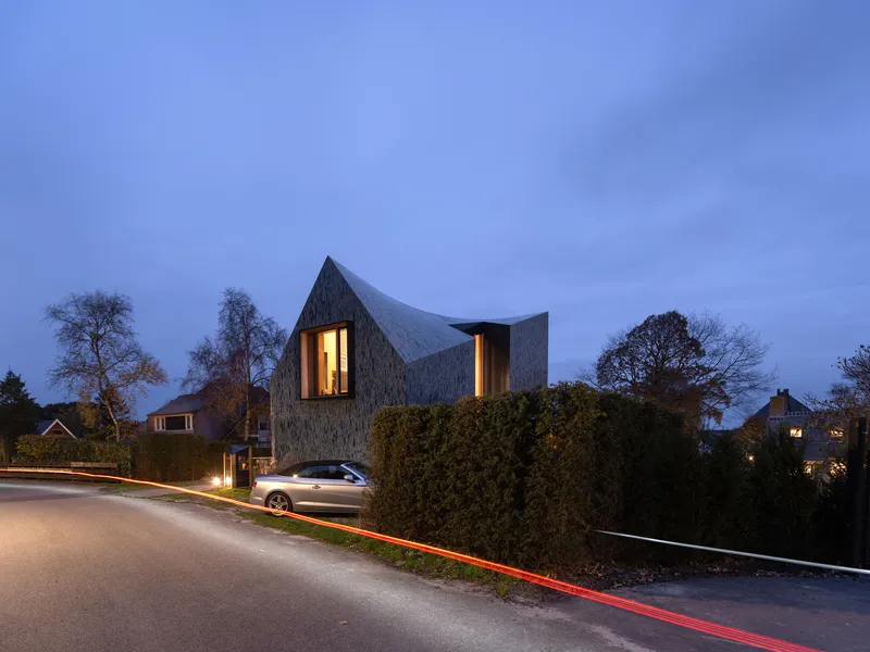 Street view of the textured facade with illuminated vertical windows at dusk behind a hedge