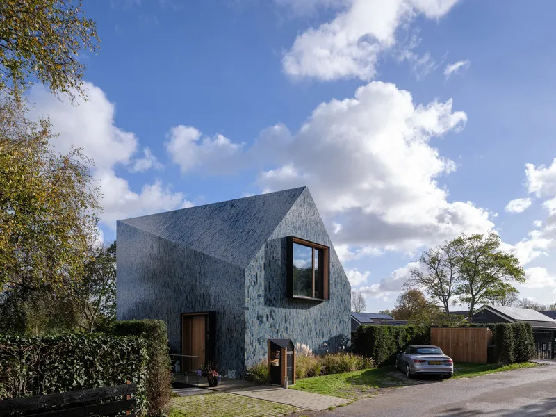 Street view of the gabled house clad in variegated blue ceramic tiles beneath white clouds