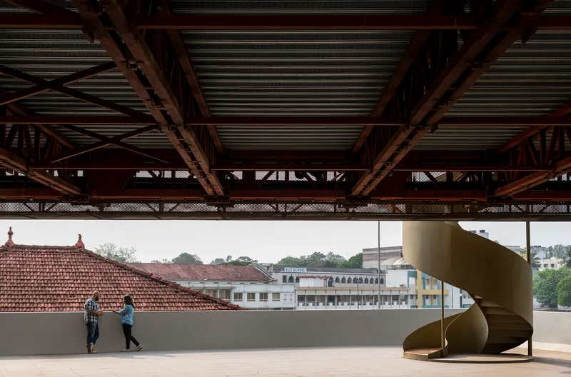 Rooftop terrace with a helical metal staircase and two figures walking past exposed trusses overhead