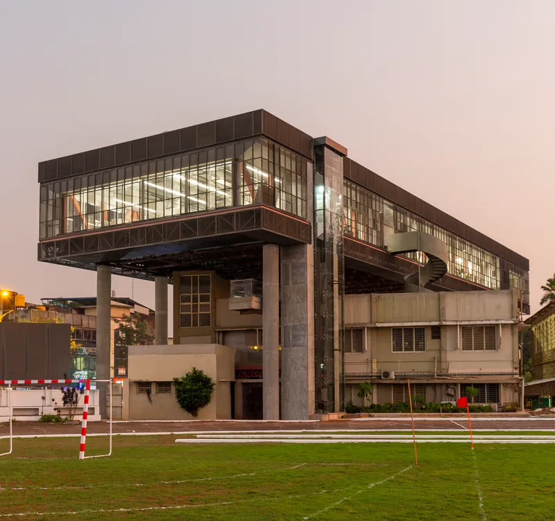 Elevated glass-wrapped volume on pilotis above existing low-rise structure at dusk